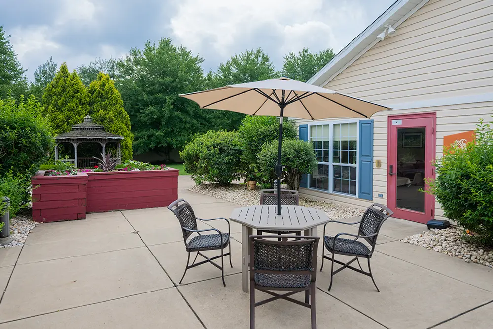 Monroeville-11 A patio with a round table, four chairs, and a large umbrella, surrounded by greenery and shrubs, next to a building with blue window shutters and a red door.