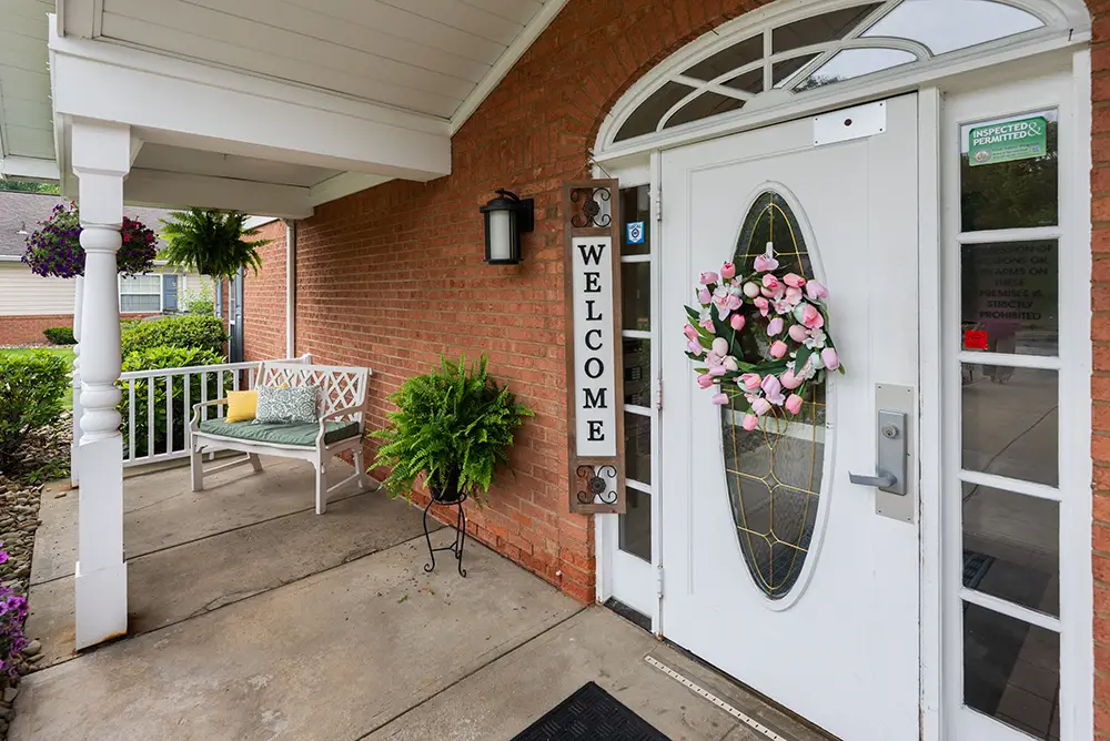 Monroeville-12 White door with oval glass and pink flower wreath, "Welcome" sign, bench with yellow cushion, and potted plants by brick exterior entrance.
