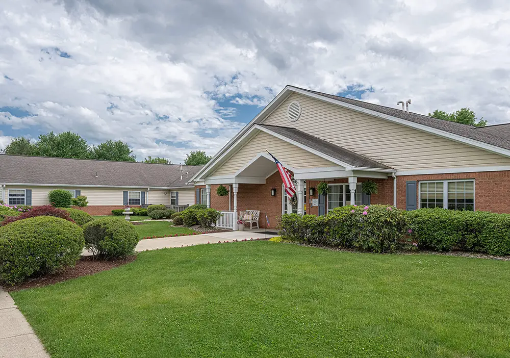 Single-story brick building in Monroeville with an American flag by the entrance, surrounded by neatly trimmed bushes and a well-kept green lawn under a cloudy sky—an inviting setting for memory care at Arden Courts.