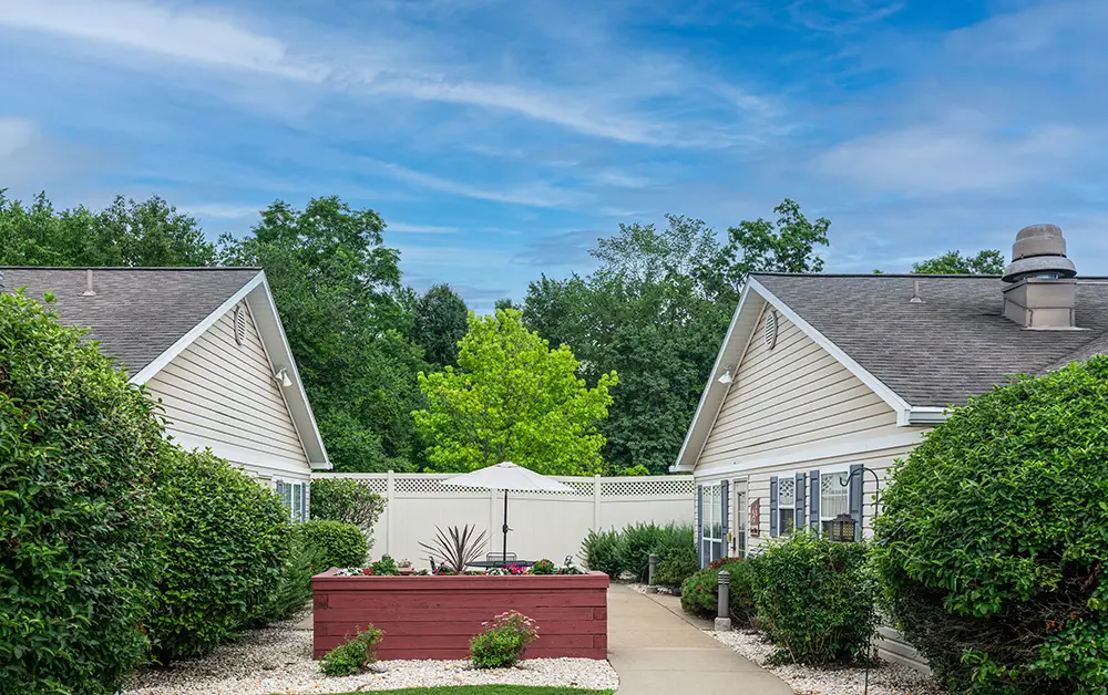 Monroeville-2 Two light-colored single-story buildings are separated by a walkway, with hedges, a planter, and a white fence at the end. Green trees and a blue sky are visible in the background.