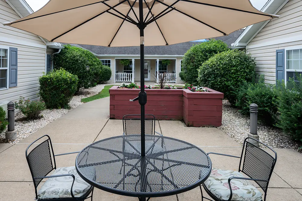 Monroeville-4 A round metal patio table with four chairs sits under a large umbrella in a courtyard with landscaped bushes and raised flower beds between two beige buildings.