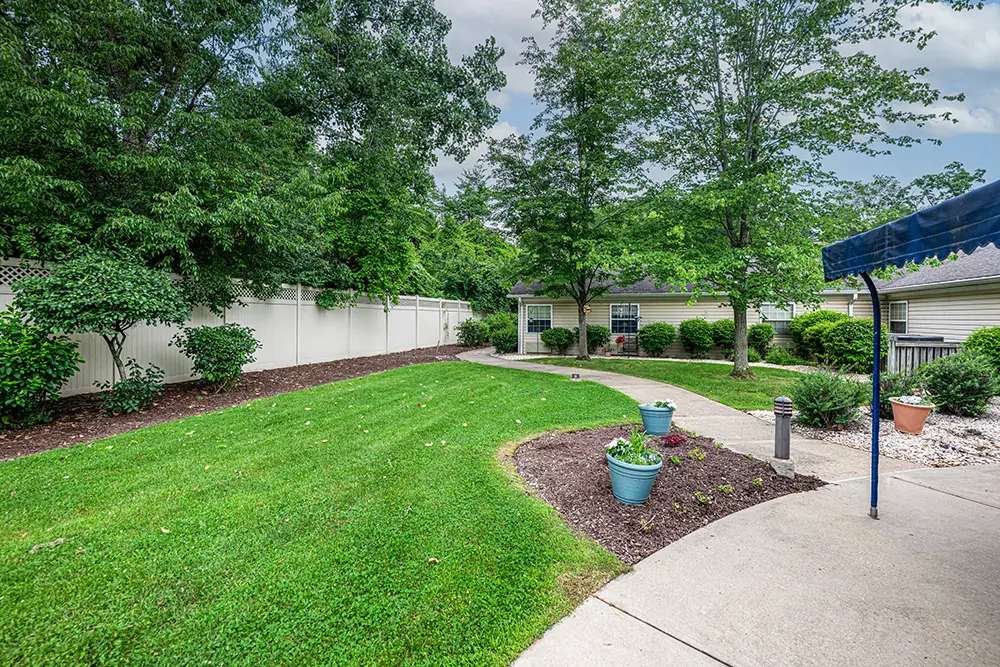 Monroeville-5 A landscaped backyard with a curved concrete path, green lawn, potted plants, and trees, bordered by a white fence and a small building in the background.