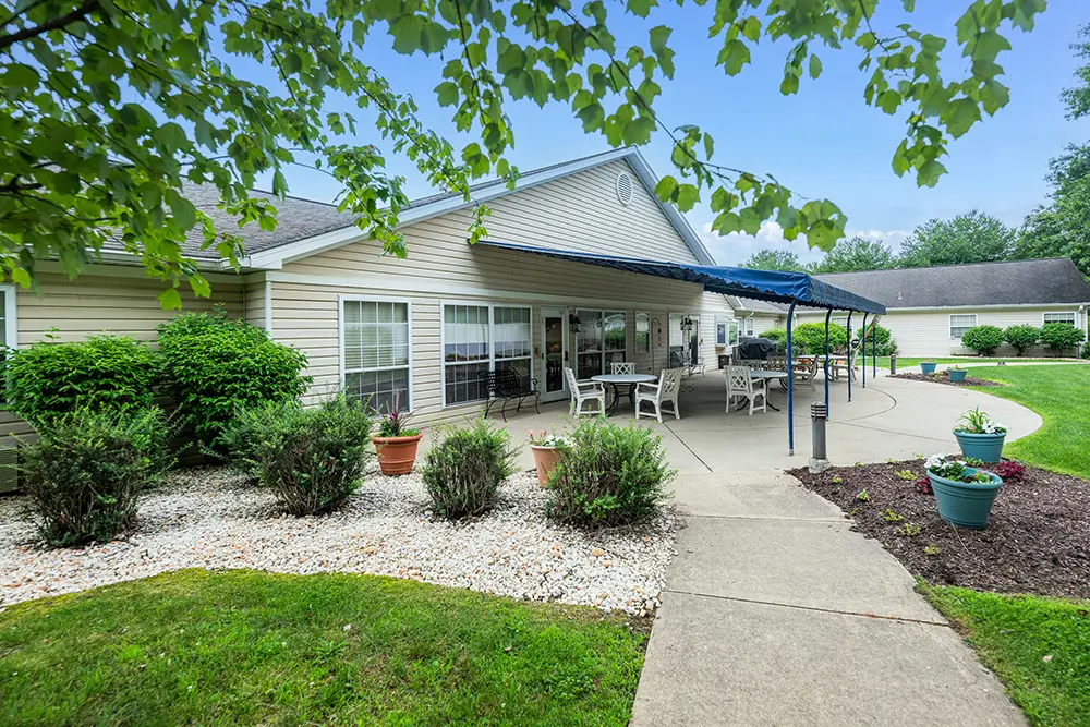 Monroeville-6 Single-story house with a covered patio area, outdoor seating, potted plants, shrubs, and a curved walkway bordered by grass and mulch.