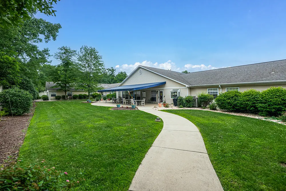 Monroeville-8 Single-story building with a covered patio and white rocking chairs, surrounded by a landscaped lawn, bushes, and a curved sidewalk under a clear blue sky.