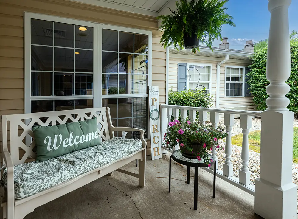 Monroeville Small front porch with a white bench, "Welcome" pillow, side table with potted flowers, hanging fern, and decorative "Porch" sign near a window.