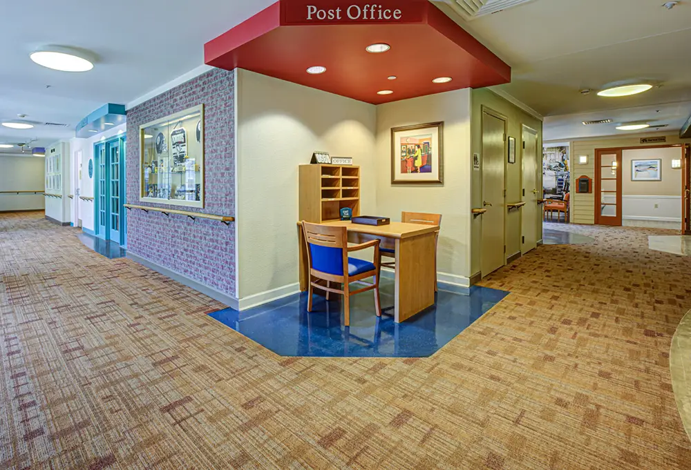 Small post office area with a desk, chair, and shelves sits in a hallway with carpeted floors and framed wall art in a public or residential building.
