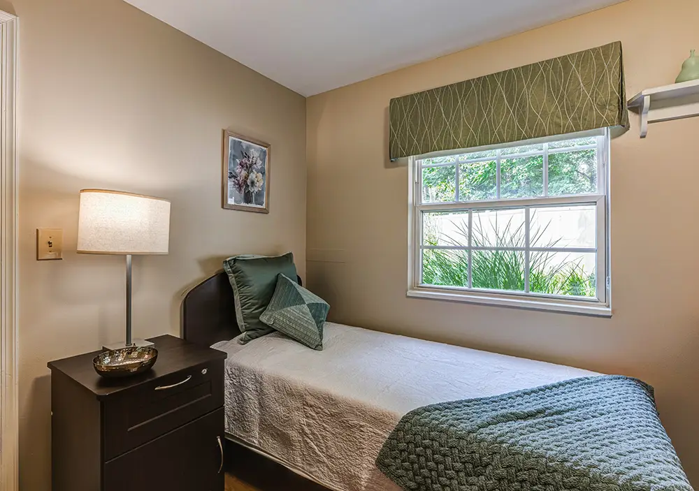 New Towson Exports03 A small bedroom with a single bed, green bedding, a nightstand with a lamp, a window with green valance, and a framed picture on the wall.