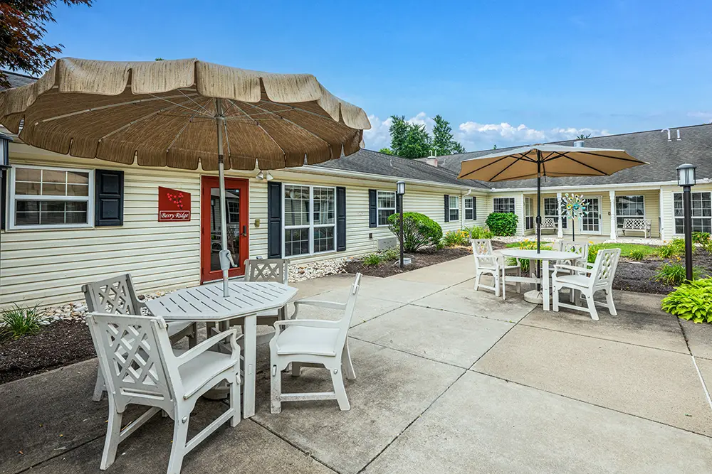 North Hills-10 Outdoor courtyard with white patio tables and chairs, large beige umbrellas, concrete walkway, landscaped plants, and beige residential building in the background.