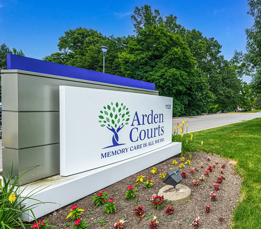 North Hills-13 Large outdoor sign for Arden Courts, a memory care facility, surrounded by flowers and greenery with a road and trees in the background.