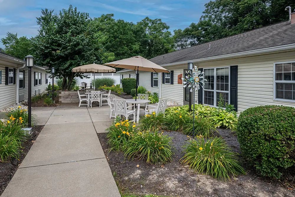 North Hills-15 Outdoor courtyard with a concrete walkway, patio tables and chairs under umbrellas, landscaped flower beds, and two single-story buildings on either side.