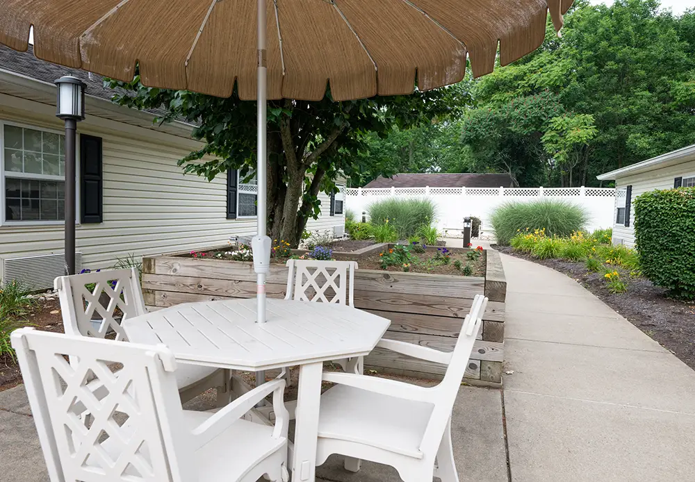 North Hills-4 A white patio table with four chairs sits under a large umbrella beside a garden bed, next to a concrete walkway and single-story building.