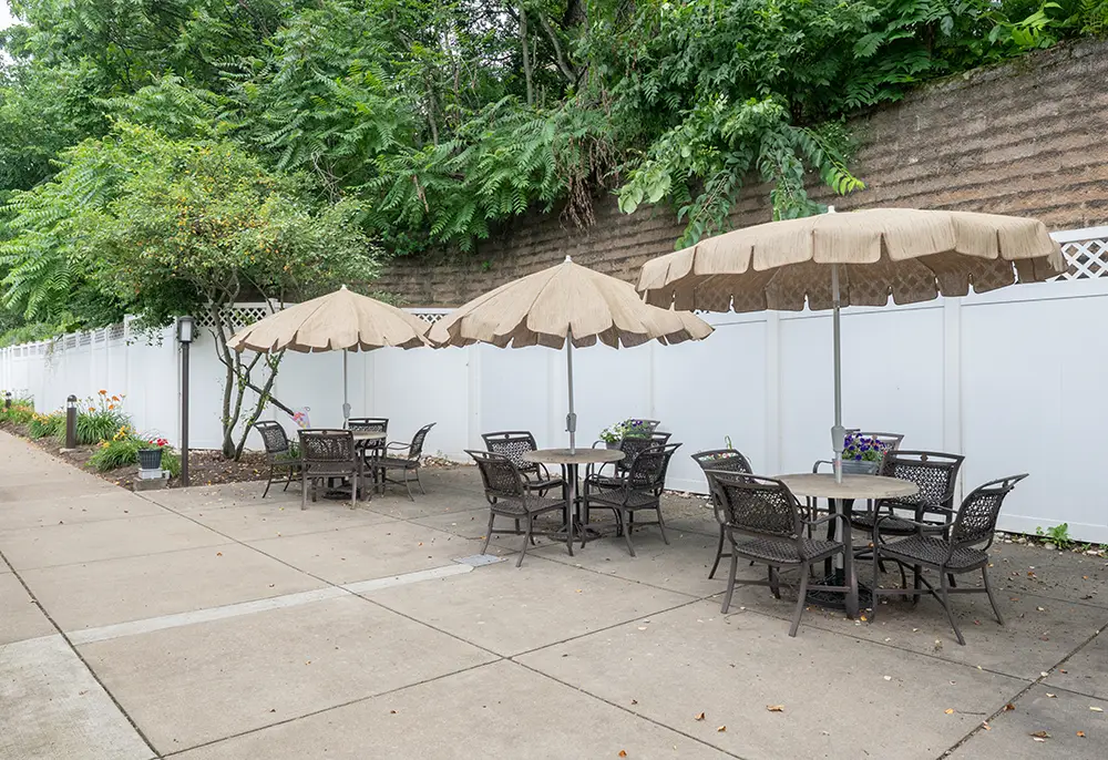North Hills-7 Outdoor patio area with three round tables, each surrounded by four chairs and shaded by beige umbrellas, next to a white fence and greenery.