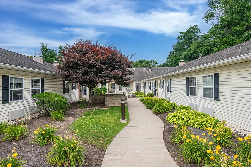 North Hills-8 Single-story building with beige siding, black shutters, and landscaped walkway lined with bushes, flowers, and a large tree in the center under a blue sky.