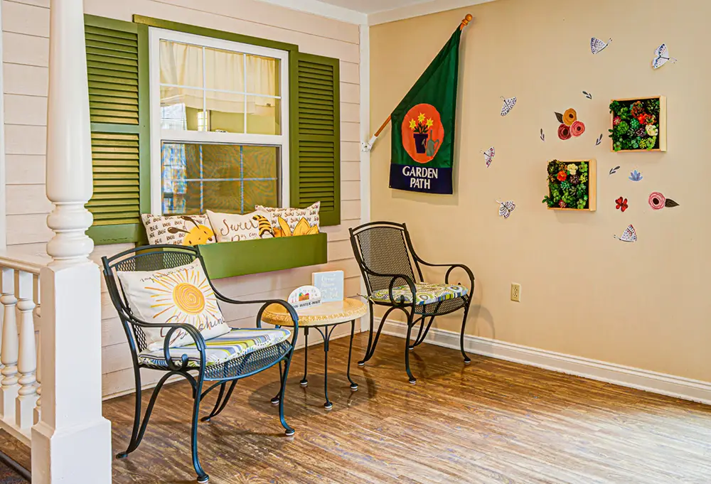 Two metal chairs with cushions and a small table are arranged in a cozy indoor nook with a decorative flag, wall art, and a window with green shutters.