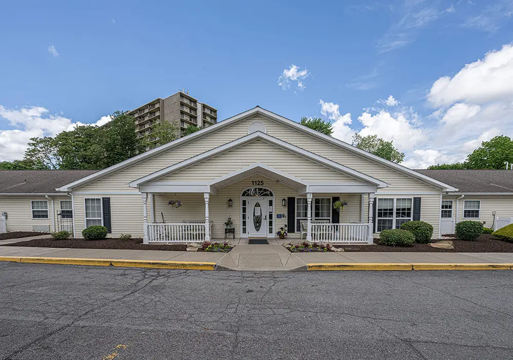 Single-story beige building with a covered entrance, white trim, and neatly landscaped shrubs; Arden Courts North Hills offers specialized memory care. A tall apartment building rises in the background under a partly cloudy sky.