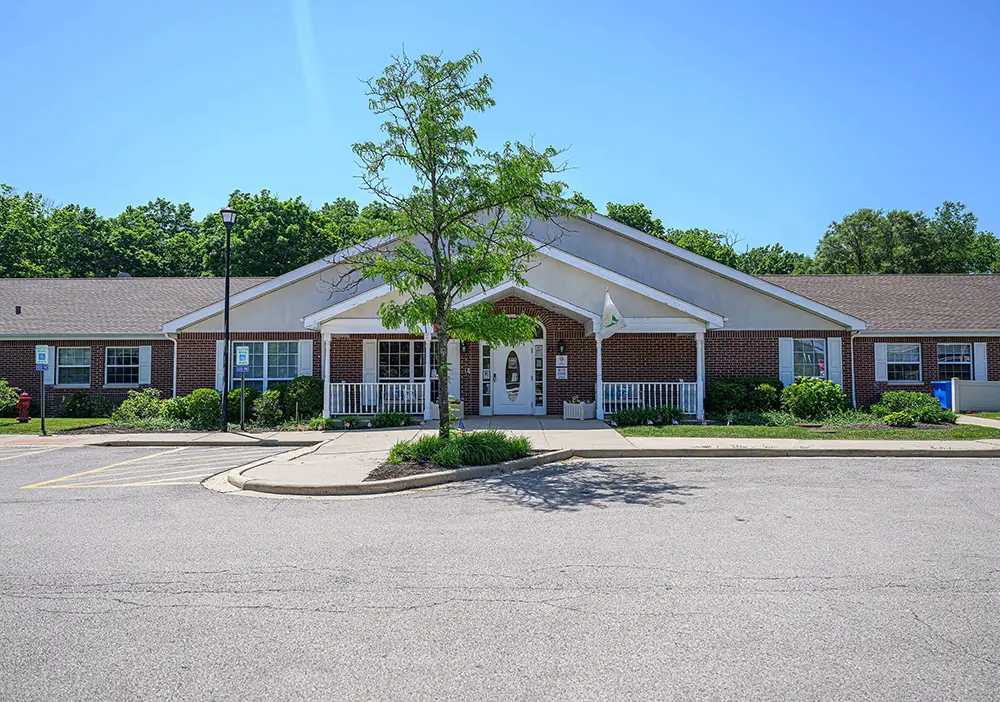 Single-story brick building with white trim, a central entrance, and covered porch in Northbrook—Arden Courts offers specialized memory care, set amid a tree and clear blue sky.
