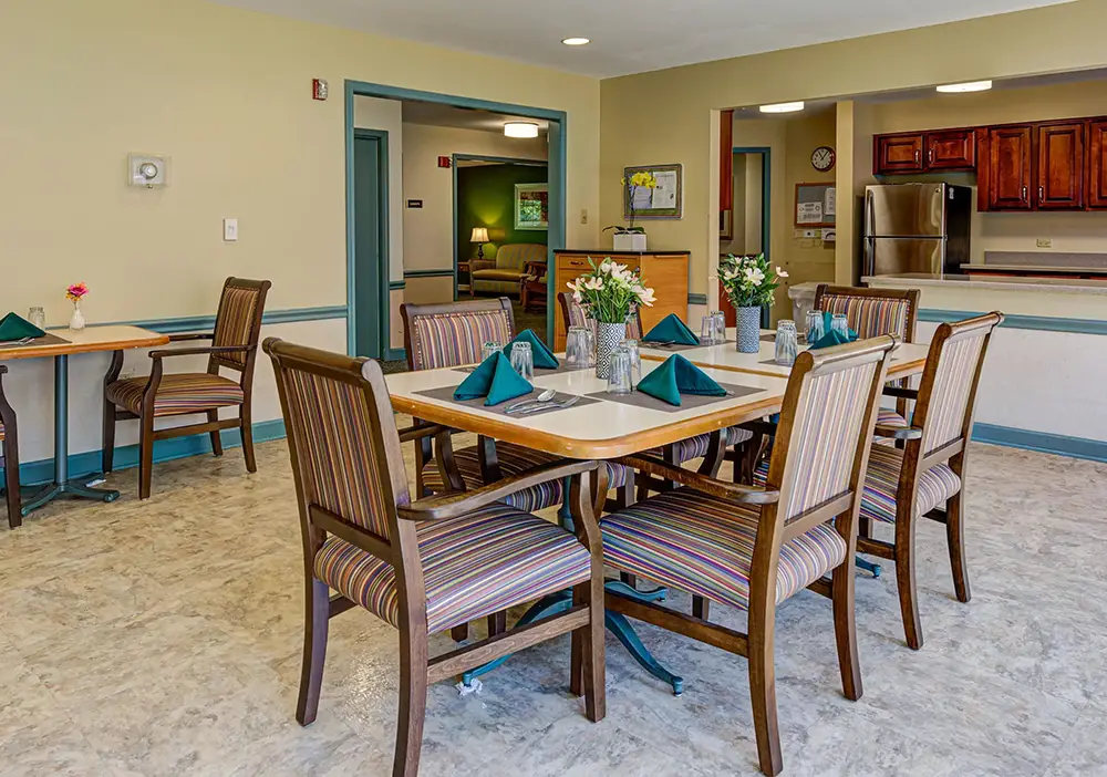 A small dining area with a table set for four, green napkins, glassware, wooden chairs, and flower centerpieces; kitchen and lounge visible in the background. A small dining area with a table set for four, green napkins, glassware, wooden chairs, and flower centerpieces; kitchen and lounge visible in the background.
