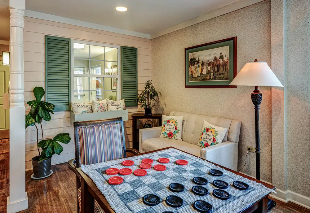A cozy living room with a checkers game on a table, a beige sofa with floral pillows, a floor lamp, a potted plant, and a framed painting on the wall.