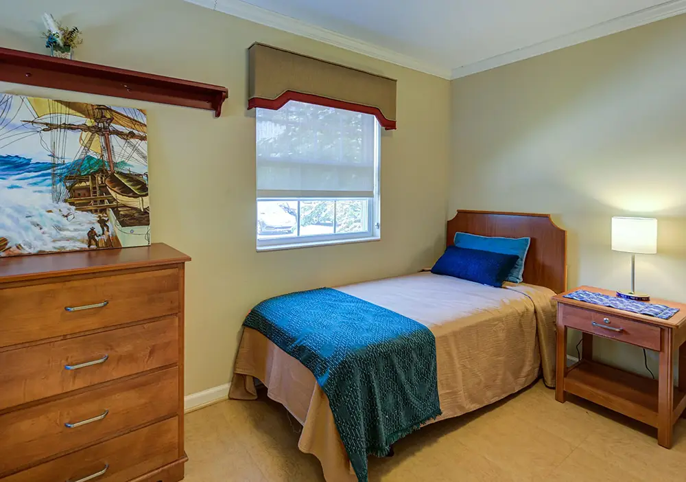 A small bedroom with a single bed, blue blanket, wooden nightstand with lamp, dresser, and a ship painting under a shelf, with natural light from a window. A small bedroom with a single bed, blue blanket, wooden nightstand with lamp, dresser, and a ship painting under a shelf, with natural light from a window.