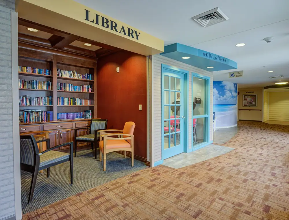 A small library nook with shelves of books, two chairs, and a table is next to a room with blue-framed glass doors in a carpeted hallway.