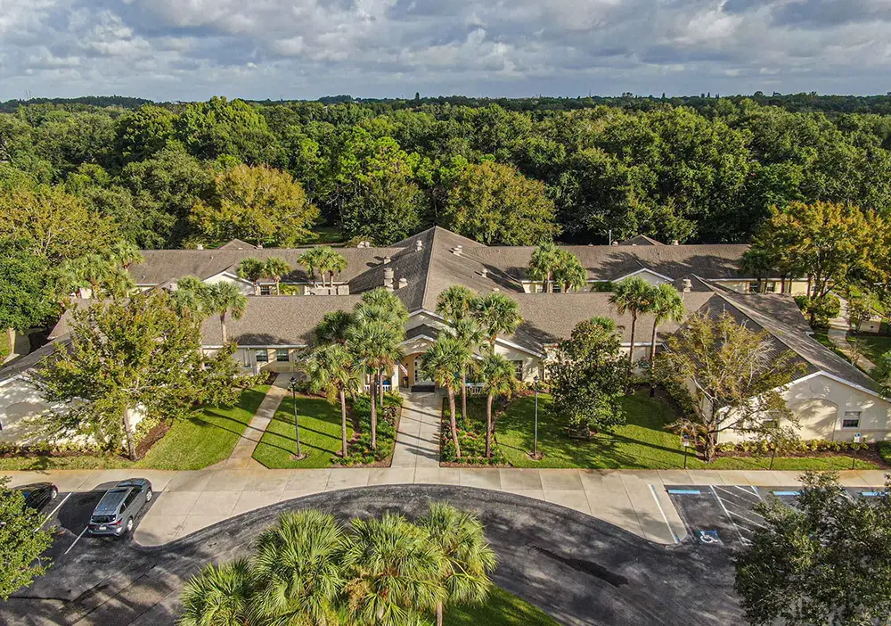 Aerial view of Arden Courts, a single-story memory care building in Palm Harbor, featuring a brown roof, surrounded by trees, landscaped lawns, and a parking area with several cars.