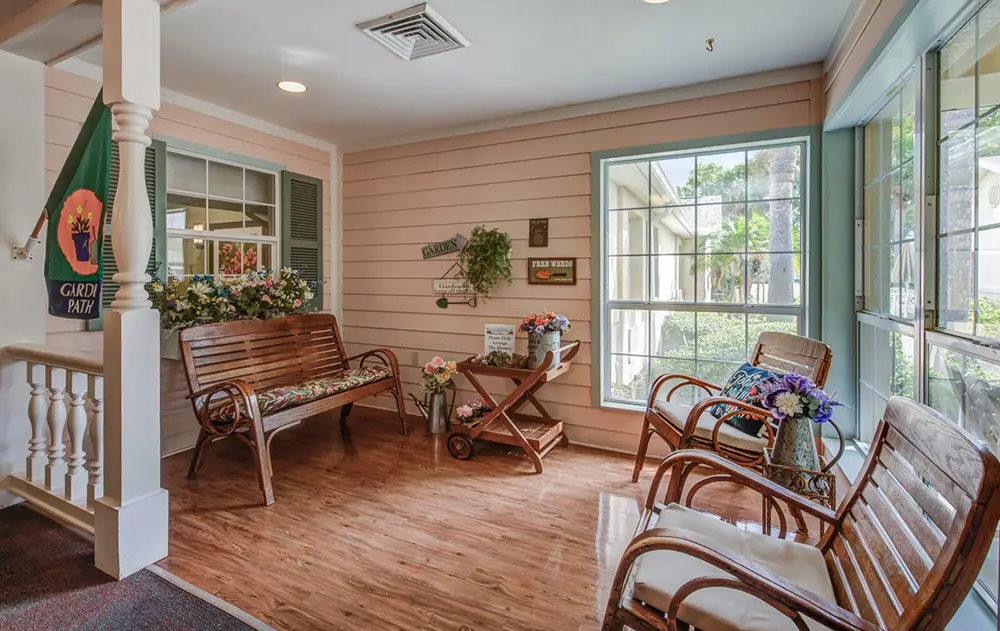 A cozy sunroom with wooden benches, chairs, a flower arrangement, wall decorations, and large windows letting in natural light.
