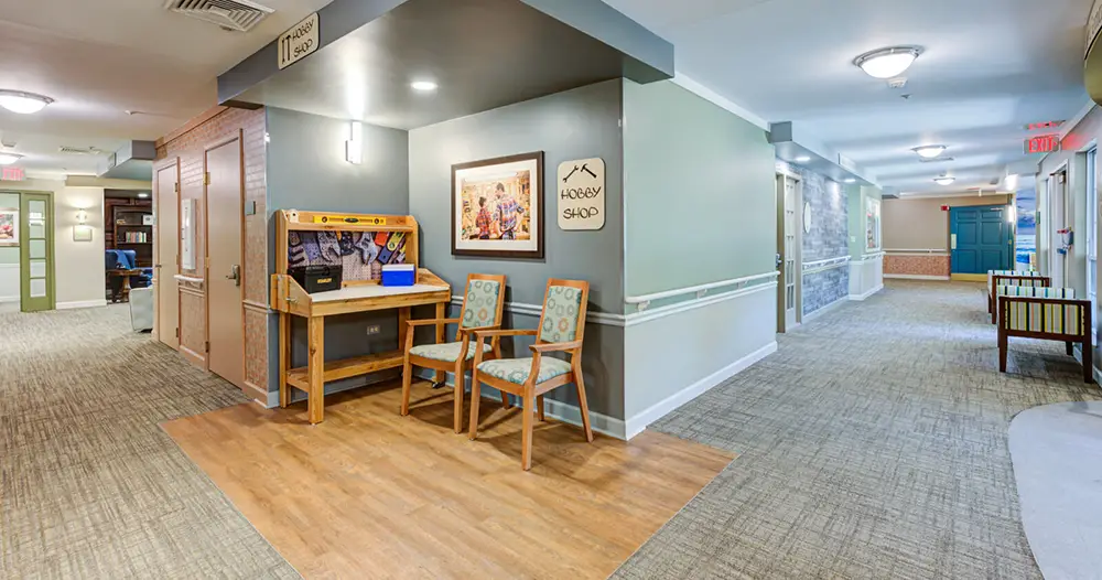 A hallway in a building features two chairs and a workbench next to a "Hobby Shop" sign, with carpeted floors and overhead lighting.