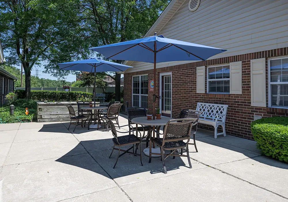 Outdoor patio at Arden Courts in Palos Heights features tables, chairs, and blue umbrellas on a concrete surface beside a brick building with windows and a white bench. Lush trees and greenery provide a serene setting for memory care residents.