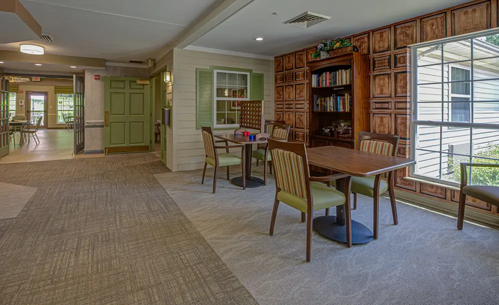 A brightly lit common area with two wooden tables, chairs, a bookshelf, large window, and view into an adjacent dining space.