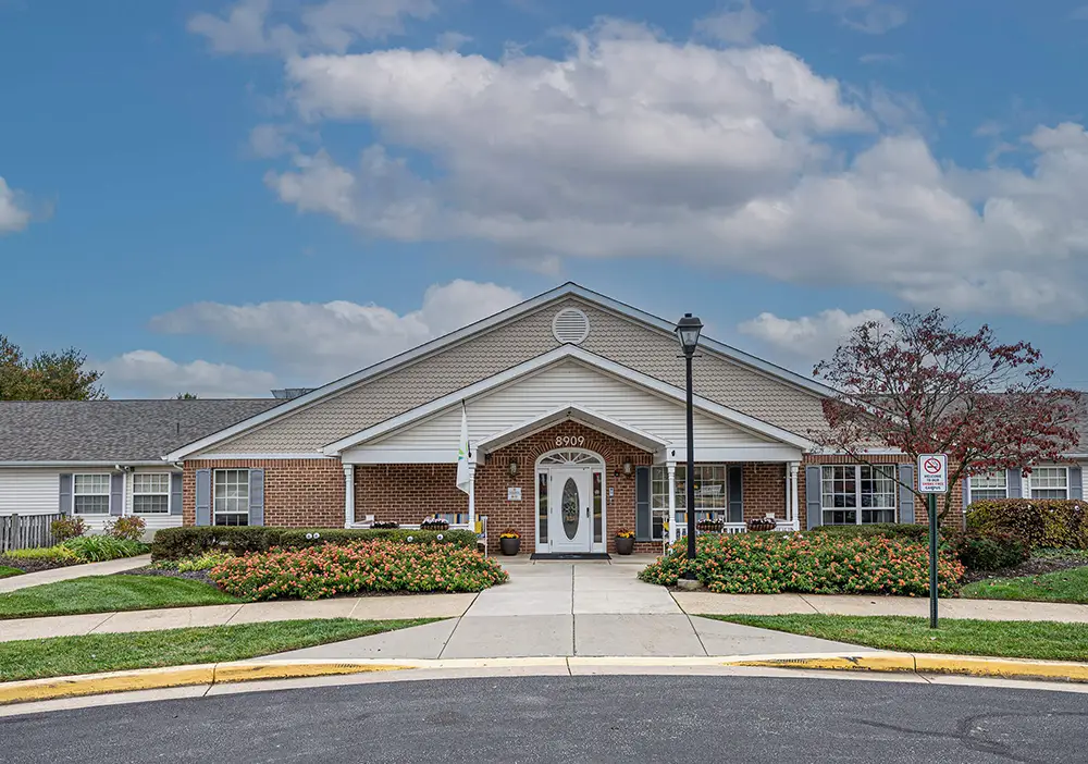 Single-story brick building with white trim, landscaped front yard, and a sidewalk leading to the entrance under a partly cloudy sky—Arden Courts in Pikesville specializes in compassionate memory care.