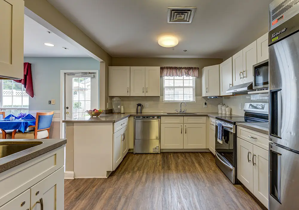 Modern kitchen with white cabinets, stainless steel appliances, wood flooring, and a window over the sink; a dining area is visible through a doorway on the left. Modern kitchen with white cabinets, stainless steel appliances, wood flooring, and a window over the sink; a dining area is visible through a doorway on the left.