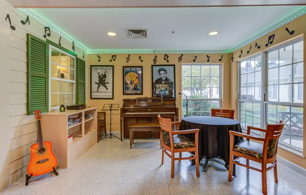 Room with a piano, guitar, round table with four chairs, wall art of musicians, bookshelves, and musical notes decorating the walls and ceiling trim. Large windows let in natural light.