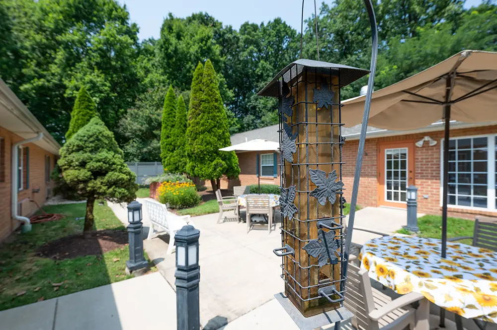 A paved patio area at Arden Courts in Potomac features outdoor tables, chairs, a bird feeder, and umbrella—perfect for memory care residents to relax among green trees and brick buildings on a sunny day. A paved patio area at Arden Courts in Potomac features outdoor tables, chairs, a bird feeder, and umbrella—perfect for memory care residents to relax among green trees and brick buildings on a sunny day.