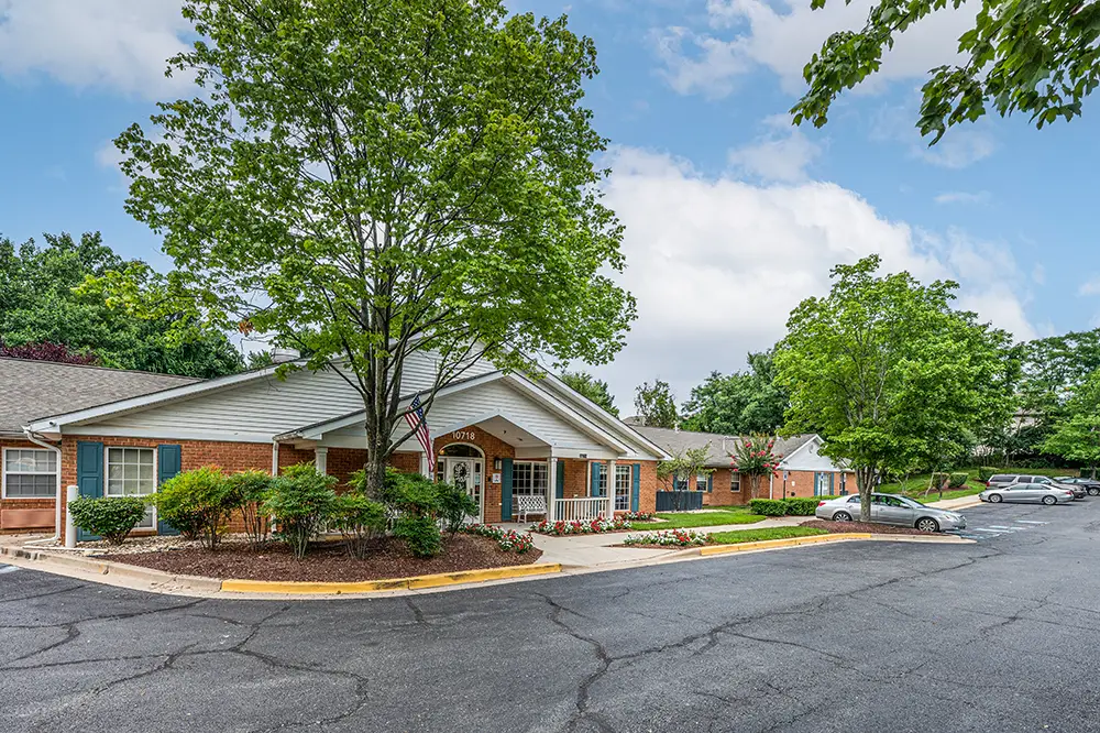 Single-story brick building with white trim, surrounded by trees and landscaping, adjacent to a parking lot under a partly cloudy sky—Arden Courts of Potomac specializes in memory care.
