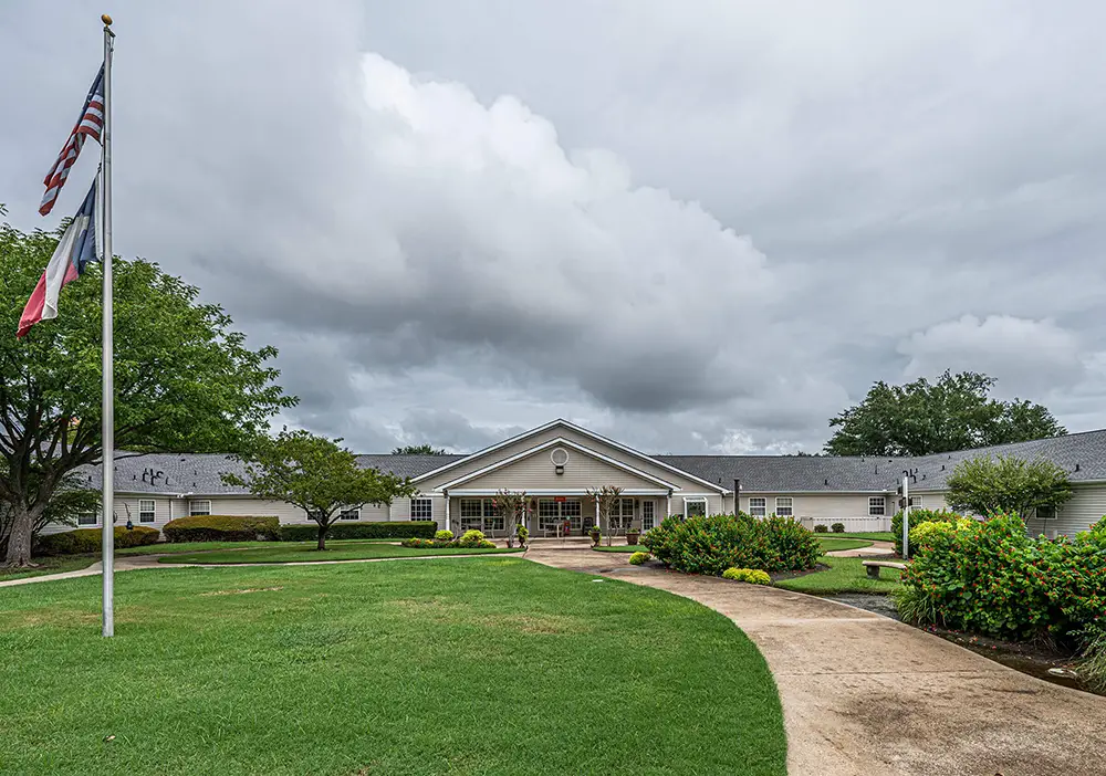 A single-story Arden Courts memory care building in Richardson features a covered entrance, surrounded by green lawns, shrubs, and trees under an overcast sky; two flagpoles stand to the left.