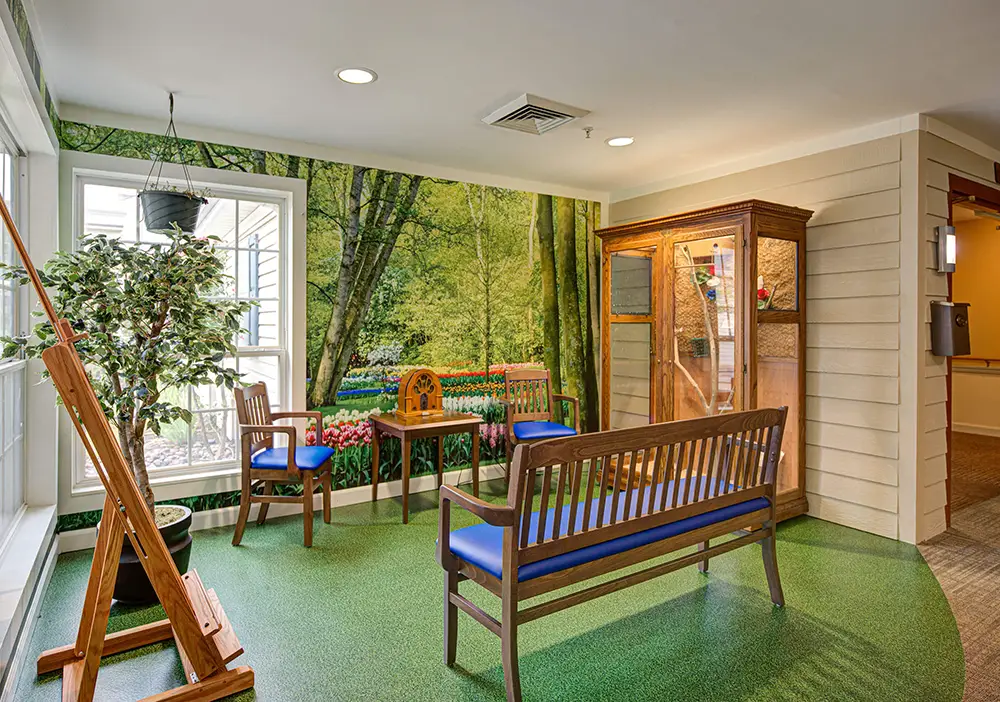 A waiting area with wooden benches, a small table, green flooring, a potted plant, a mural of a forest with flowers, and a glass-fronted wooden cabinet. A waiting area with wooden benches, a small table, green flooring, a potted plant, a mural of a forest with flowers, and a glass-fronted wooden cabinet.