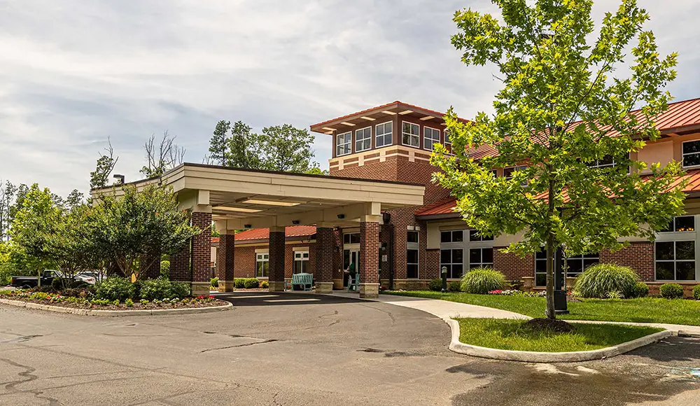 A modern brick building in Richmond with a covered entrance, landscaped greenery, and a tree in the foreground under a partly cloudy sky—perfect for memory care at Arden Courts.