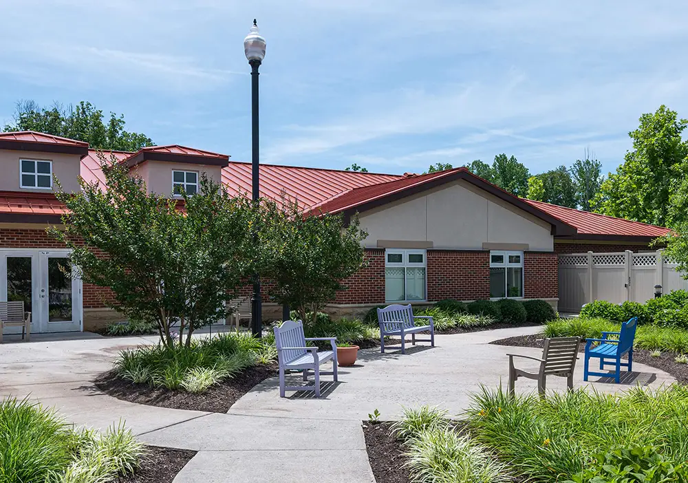 A brick building with a red roof is surrounded by shrubs and trees. Several benches and a streetlamp are on the curved walkway in the landscaped courtyard. A brick building with a red roof is surrounded by shrubs and trees. Several benches and a streetlamp are on the curved walkway in the landscaped courtyard.