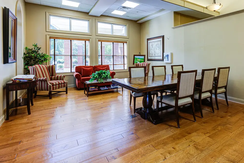 A dining area with a wooden table and eight chairs, a red sofa, two striped armchairs, potted plants, large windows, and light wood flooring.