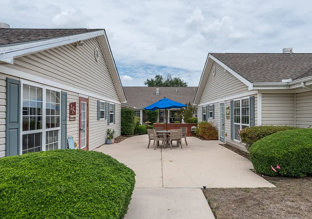 A concrete walkway at Arden Courts, a San Antonio memory care community, leads between two beige single-story buildings with white trim to a patio area featuring a table, chairs, and blue umbrella. Shrubs line the walkway.