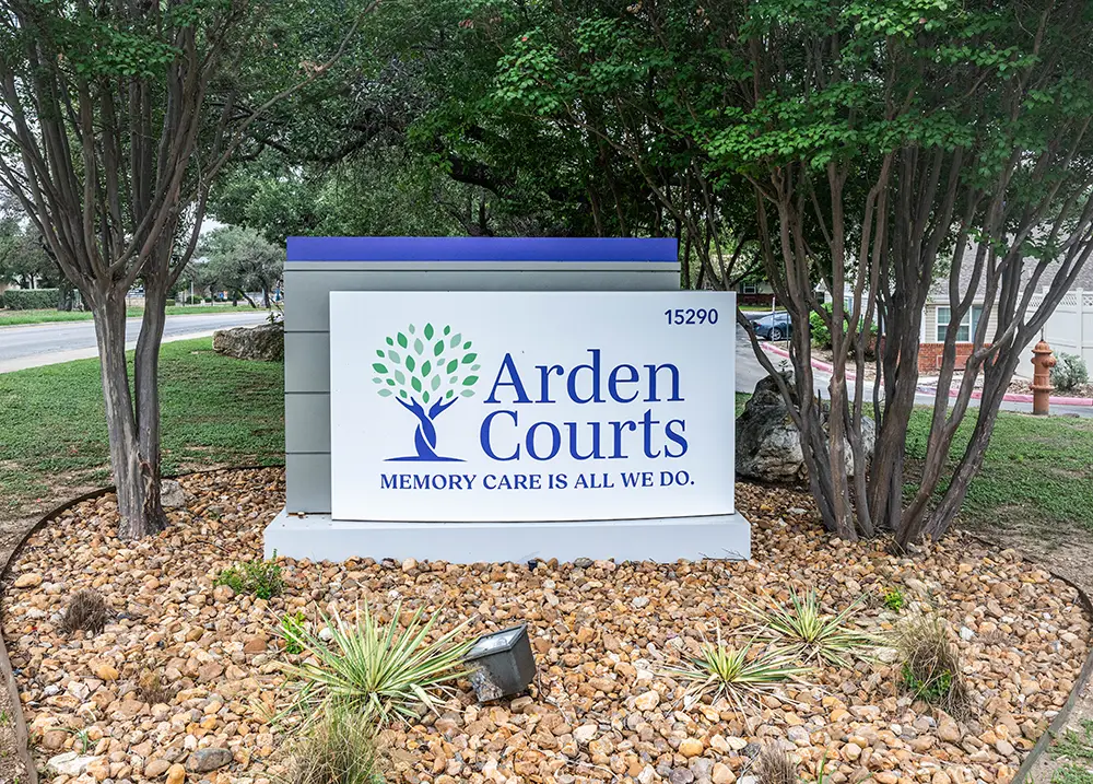 San Antonio Sign Sign for Arden Courts, a memory care facility, surrounded by rocks and plants, with trees and a road in the background.
