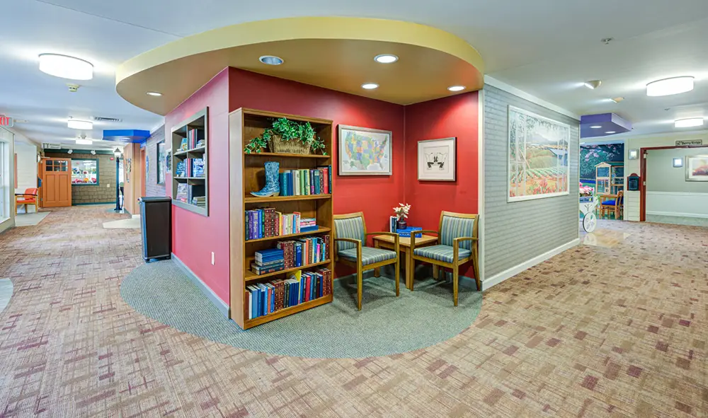 A hallway with carpeted floors, a red accent wall, a bookshelf filled with books, two striped chairs, framed art, and overhead lighting.