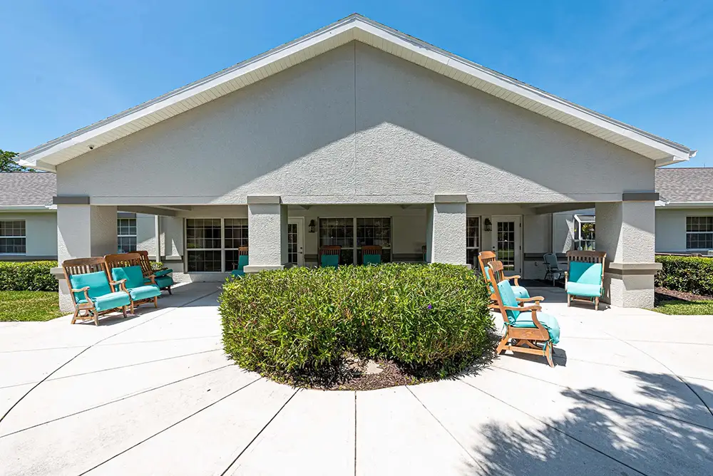 A residential building in Sarasota with a covered patio, outdoor seating, and green bushes under a clear blue sky, ideal for memory care at Arden Courts.