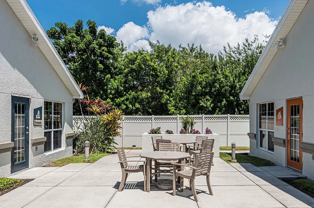 Outdoor patio area with a round table and six chairs, situated between two single-story buildings, with a white fence and trees in the background. Outdoor patio area with a round table and six chairs, situated between two single-story buildings, with a white fence and trees in the background.
