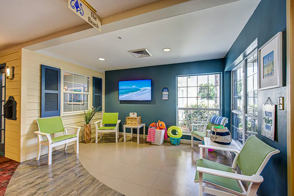 A waiting area with green cushioned chairs, beach-themed decor, a wall-mounted TV displaying an ocean scene, and large windows letting in natural light.
