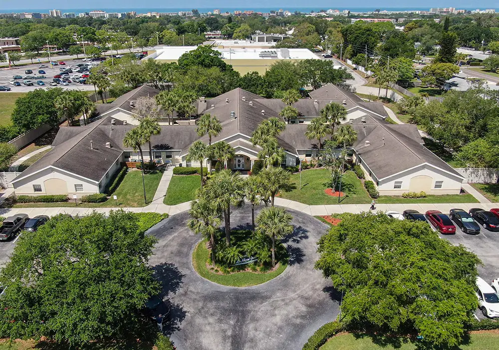 Aerial view of Arden Courts, a large, single-story building in Seminole with a circular driveway, palm trees, parked cars, and lush greenery—an inviting suburban setting dedicated to specialized memory care.