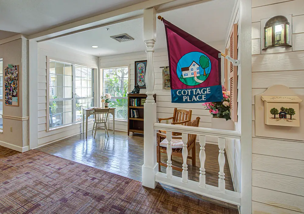 A cozy indoor area with large windows, a small desk and chair, a bookshelf, and a “Cottage Place” flag hanging by a wooden railing. A cozy indoor area with large windows, a small desk and chair, a bookshelf, and a “Cottage Place” flag hanging by a wooden railing.