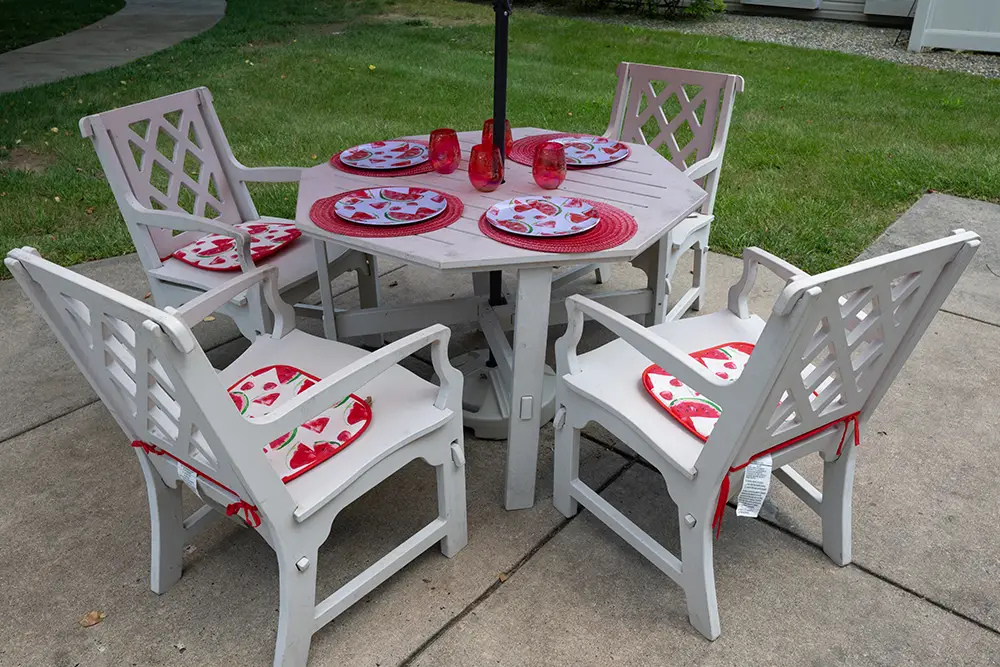 Sequence (1)Cedar Ridge-10 A round outdoor table with four white chairs, set with red placemats, red glasses, and plates featuring watermelon designs, placed on a patio near a grassy lawn.