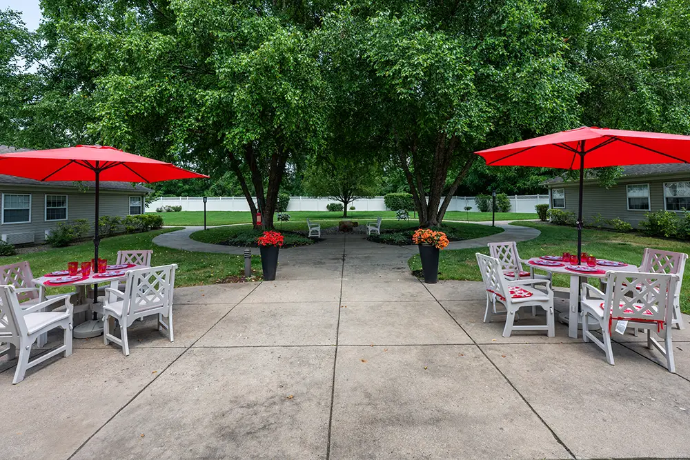 Sequence (1)Cedar Ridge-11 Outdoor patio with white tables and chairs, red umbrellas, and potted flowers; surrounded by grass, trees, and nearby buildings.