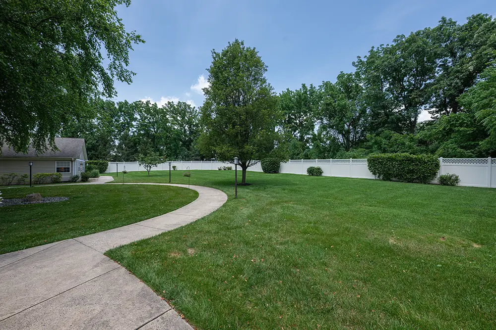 Sequence (1)Cedar Ridge-13 A curved concrete walkway runs through a large, grassy backyard with trees and a white privacy fence under a blue sky.